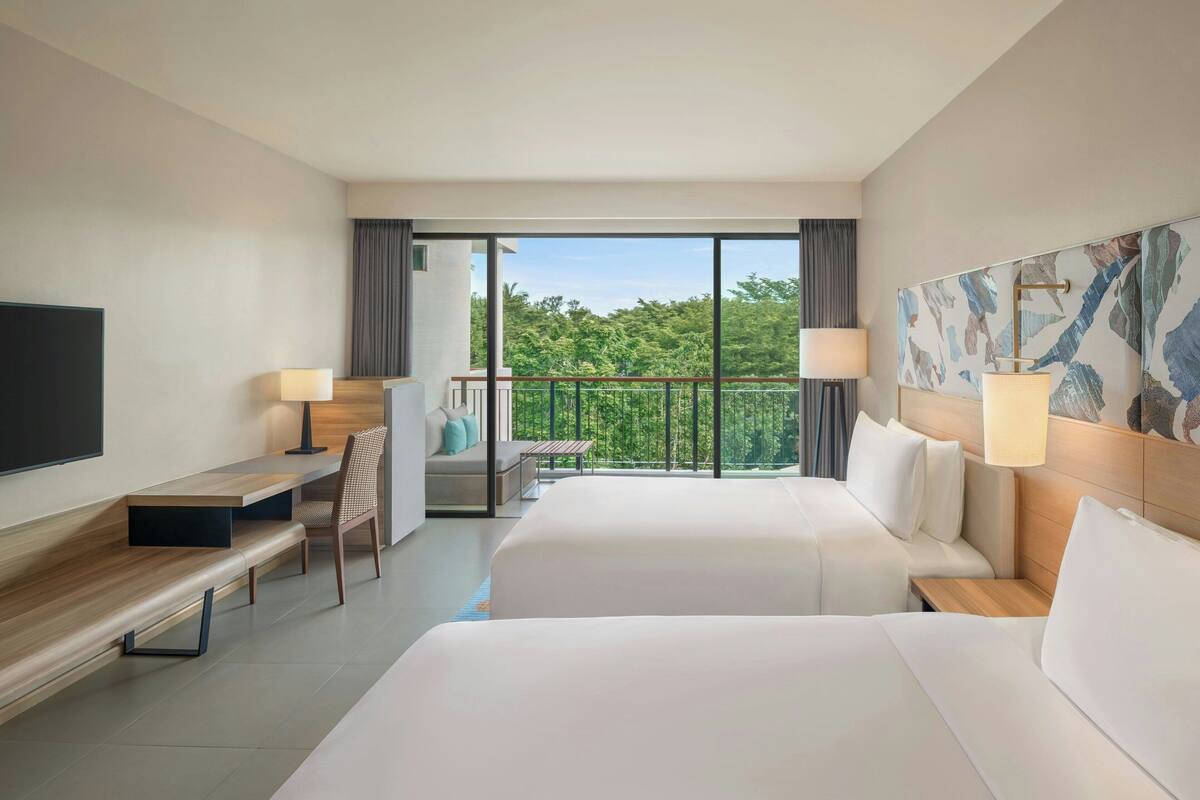 A hotel room with two beds, a desk, a chair, a TV, and a balcony with a view of greenery.