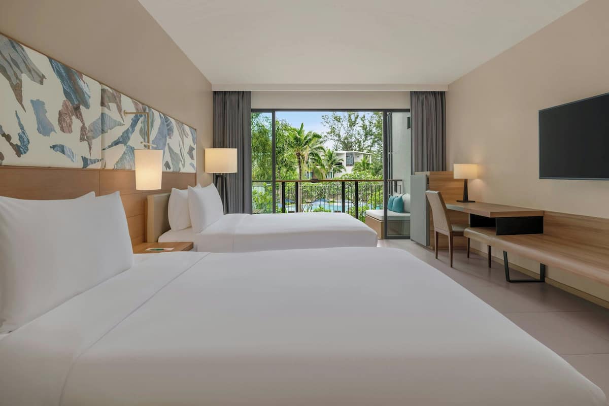 A hotel room with two beds, a desk, a chair, a TV, and a balcony with a view of palm trees and buildings.