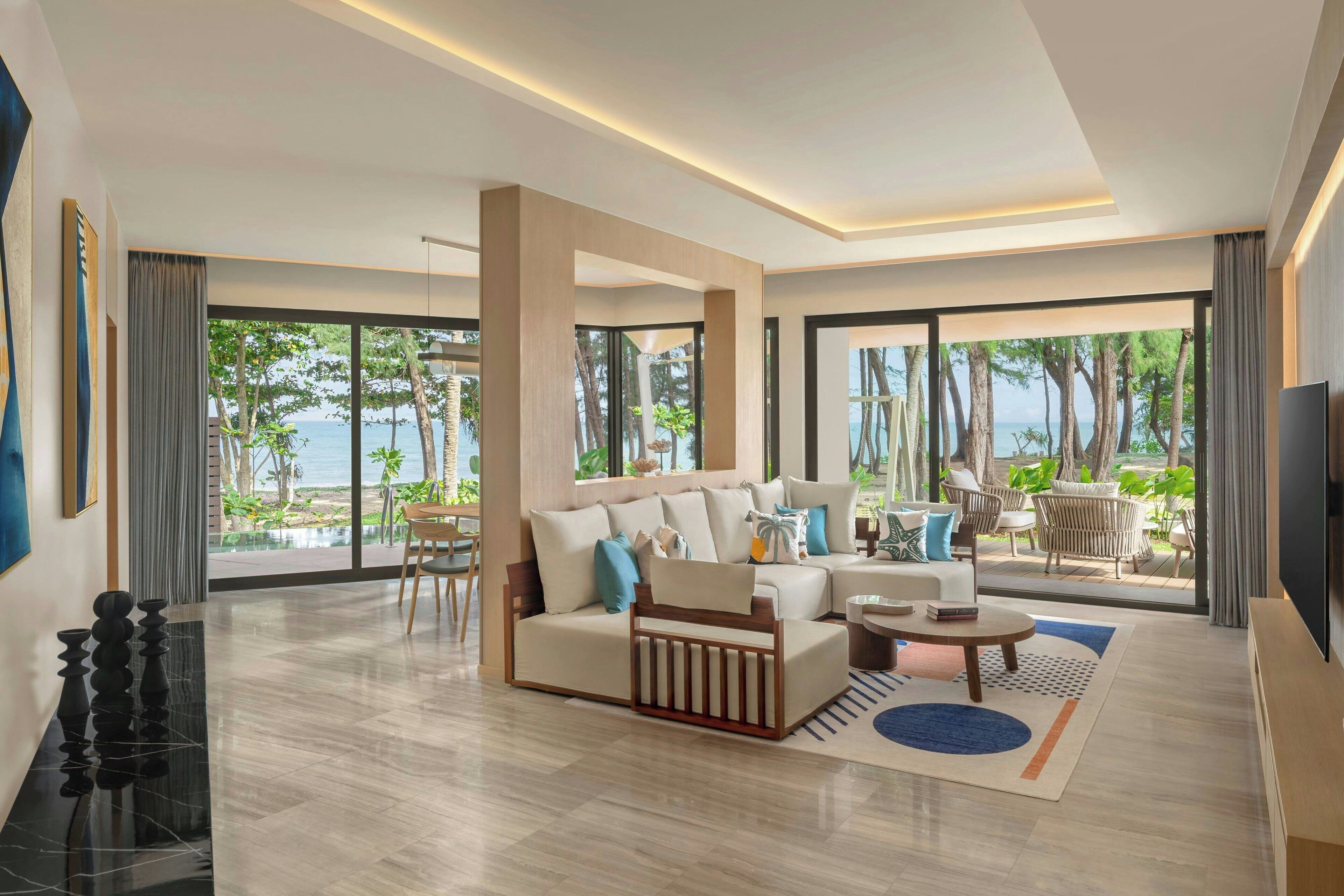 A modern living room with a sofa, coffee table, and TV, large windows with a view of the sea, and a dining area with chairs.