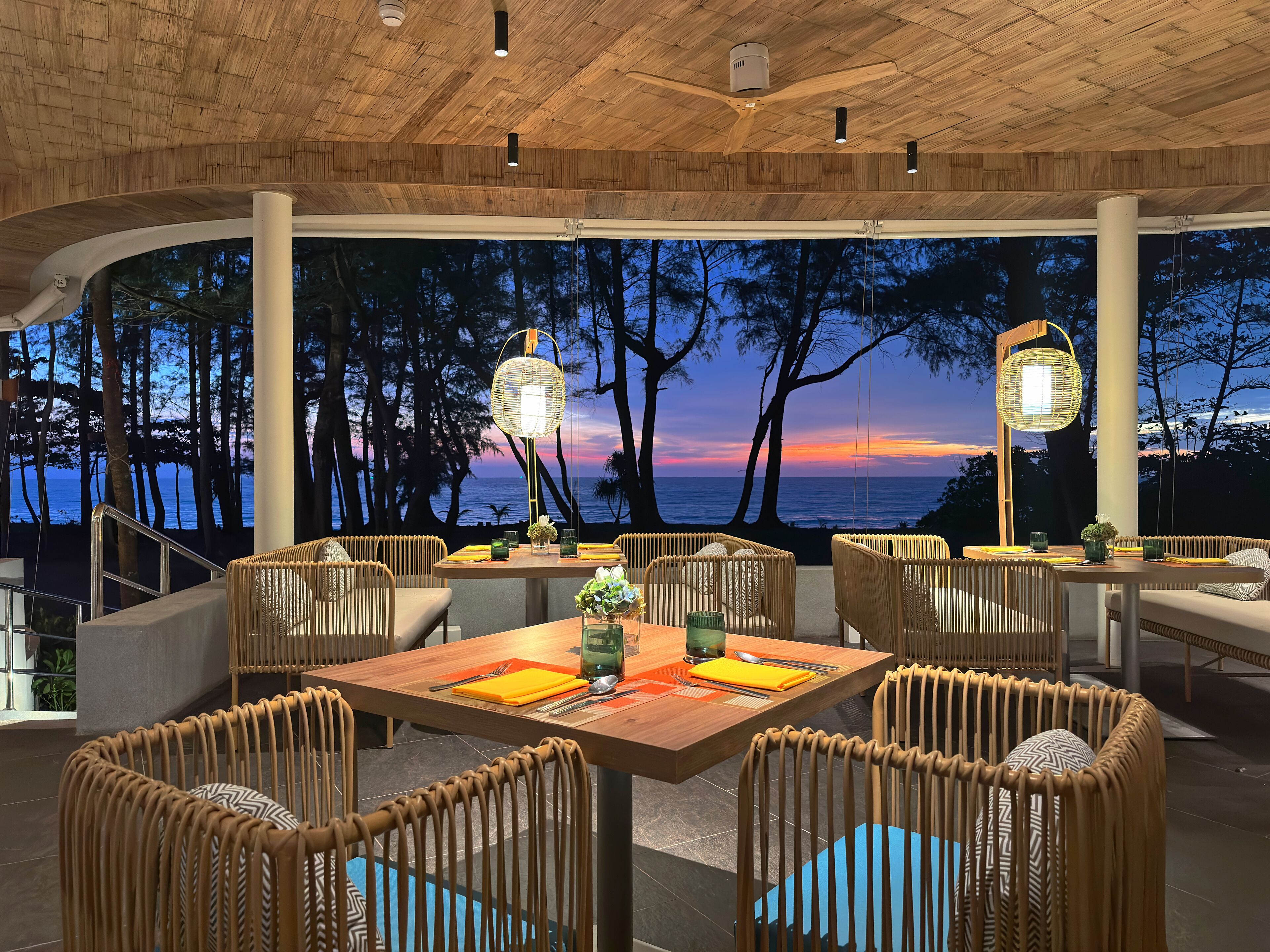 An outdoor dining area with wicker furniture, a wooden ceiling, and a view of the ocean at sunset.