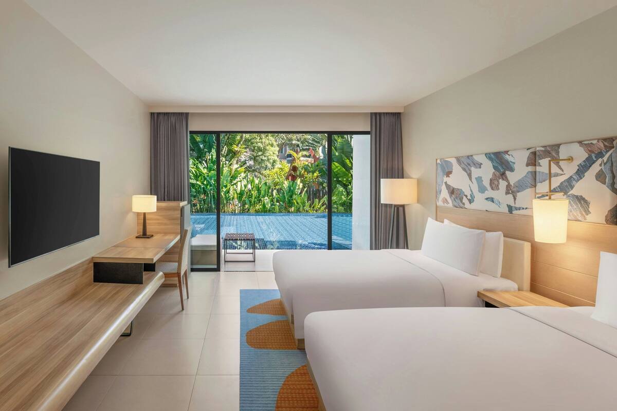 A hotel room with two beds, a TV, a desk, and a view of a swimming pool and greenery.