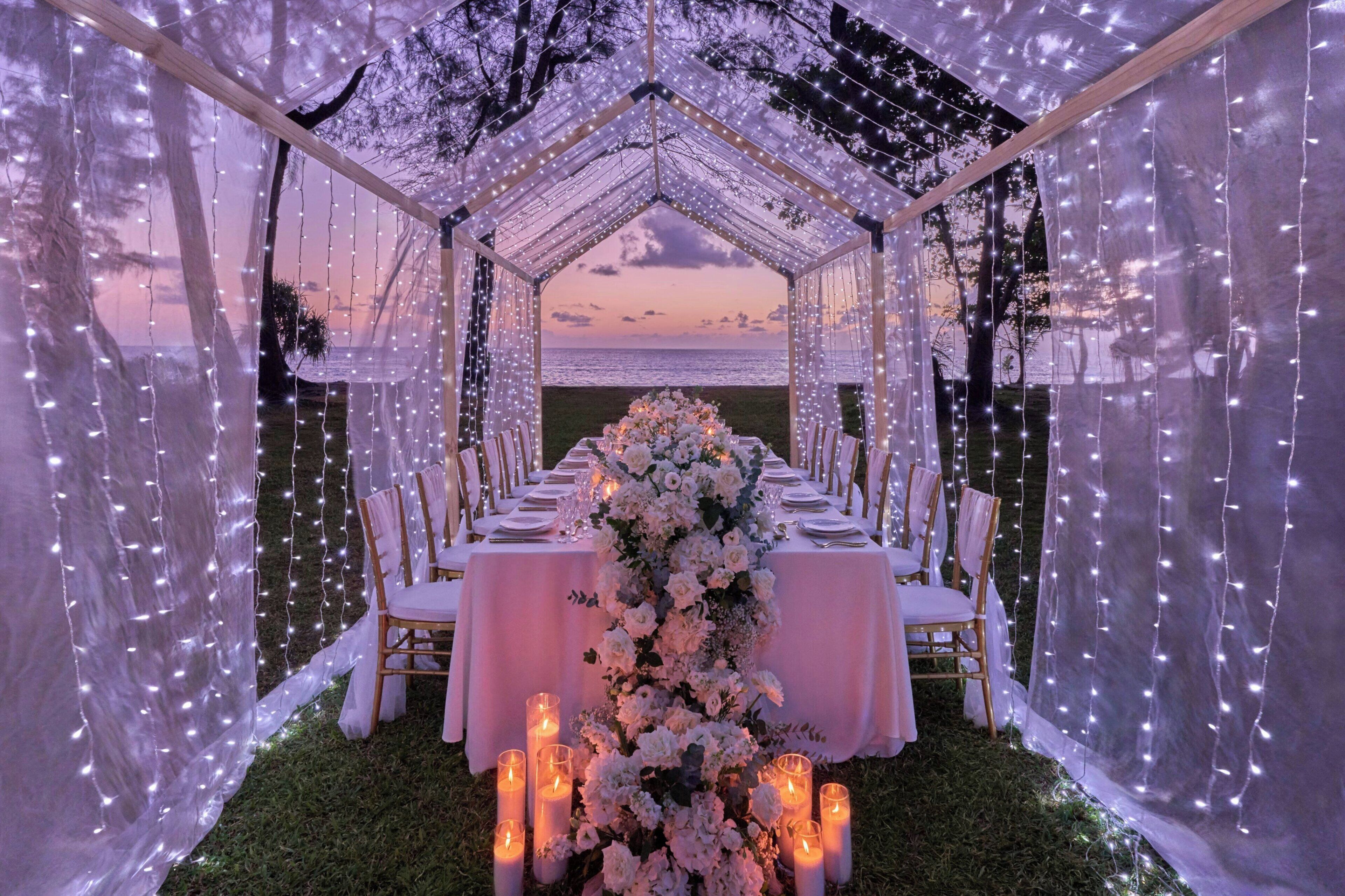 An outdoor wedding setup with a tent, white tablecloths, floral arrangements, and candles.