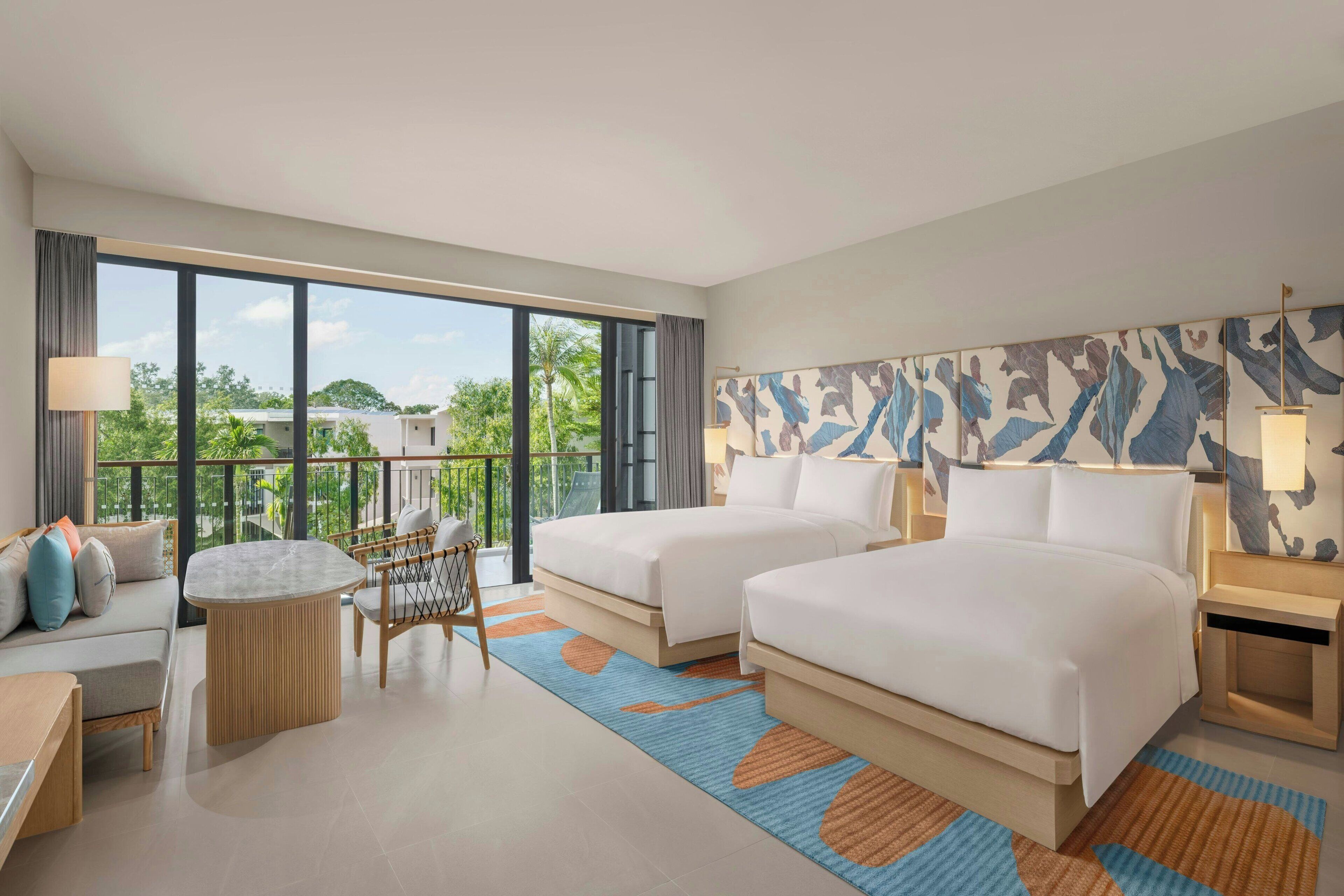 A hotel room with two beds, a sofa, a small table, and a balcony with a view of greenery.