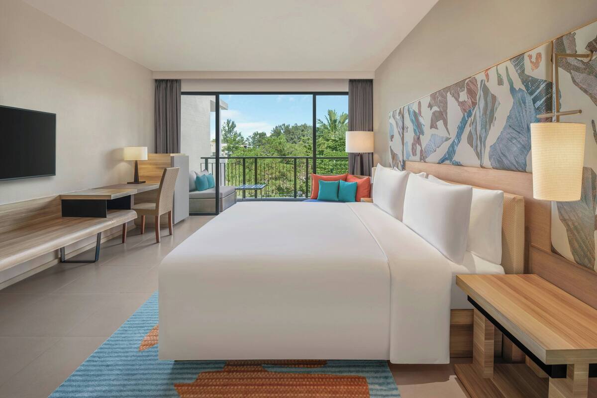 A hotel room with a large bed, a desk, a chair, a TV, and a balcony with a view of greenery.