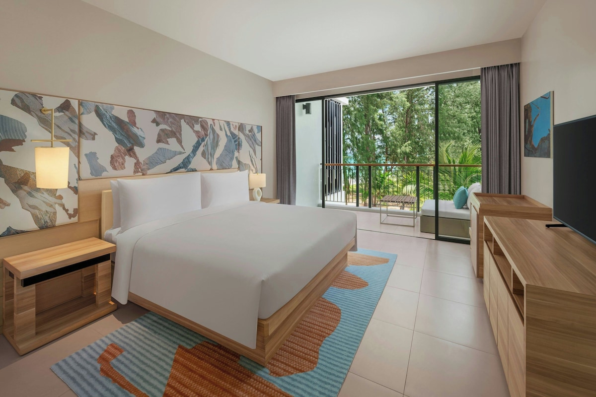 Quarto de hotel moderno com uma cama grande, televisão, escrivaninha e varanda com vista para árvores.