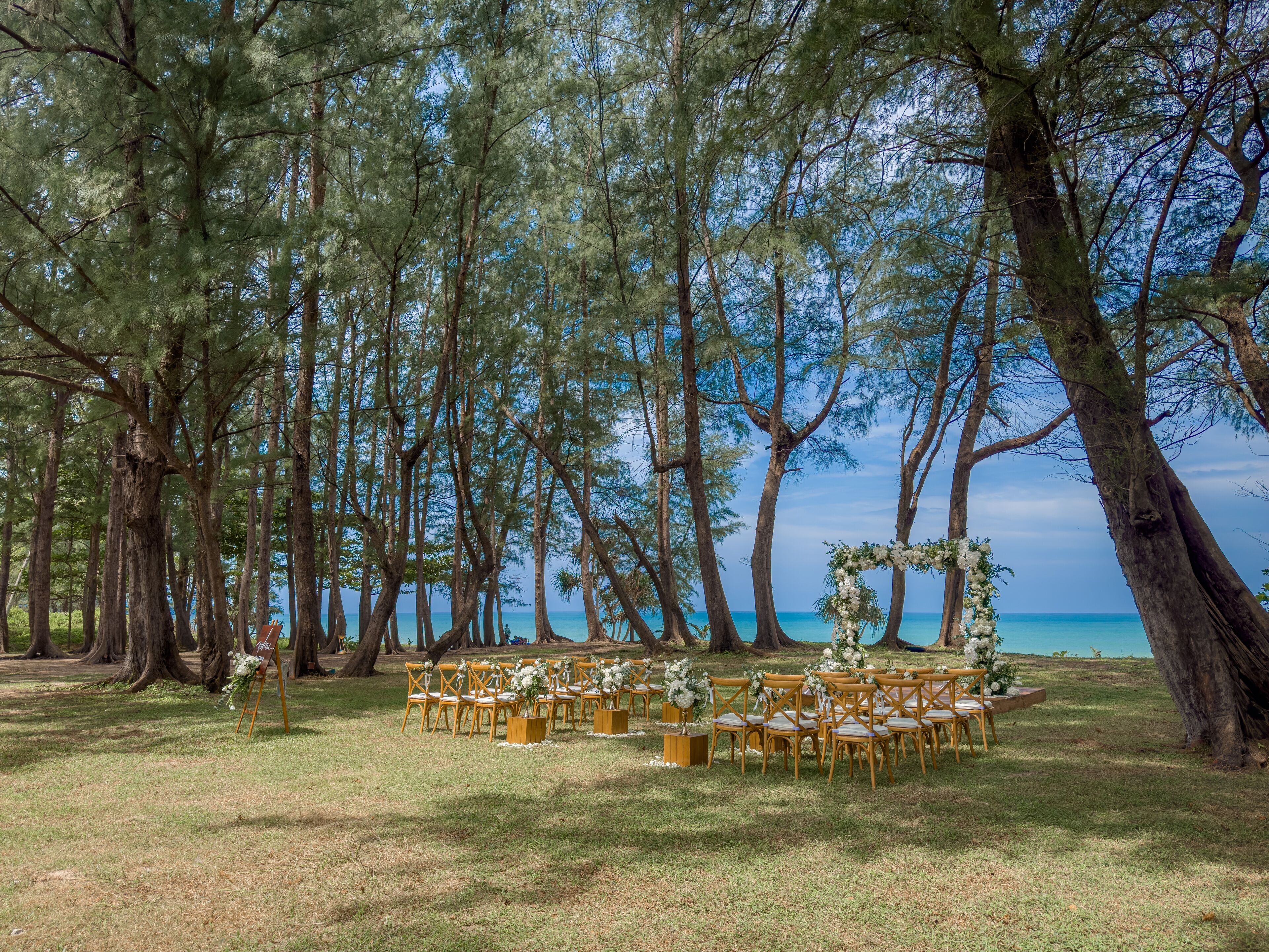 Hochzeitstisch draußen mit Holzkästen und Blumen, umgeben von hohen Bäumen und Blick auf das Meer.