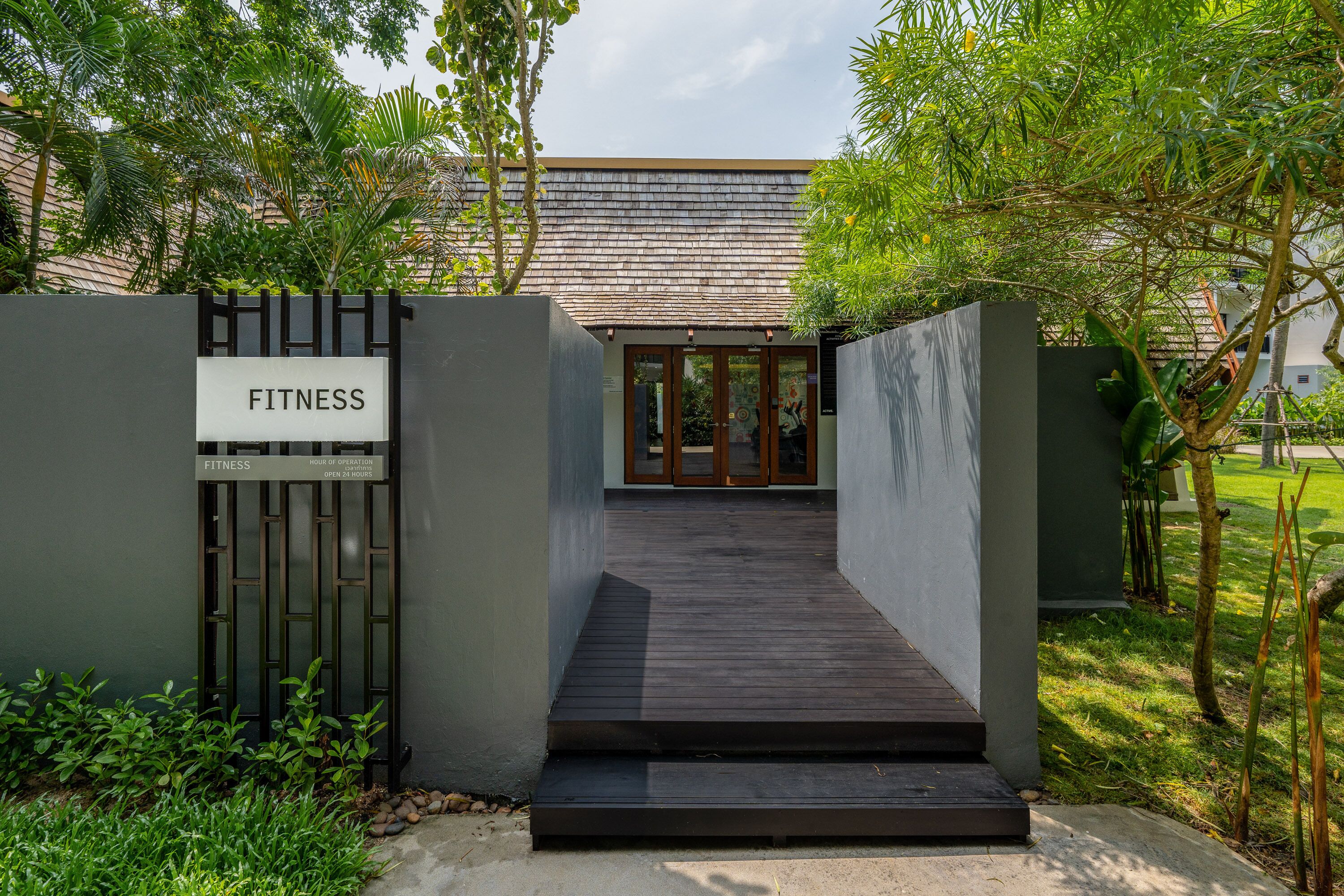 Entrada a un gimnasio con una rampa de madera que lleva a una puerta de vidrio, rodeada de follaje.