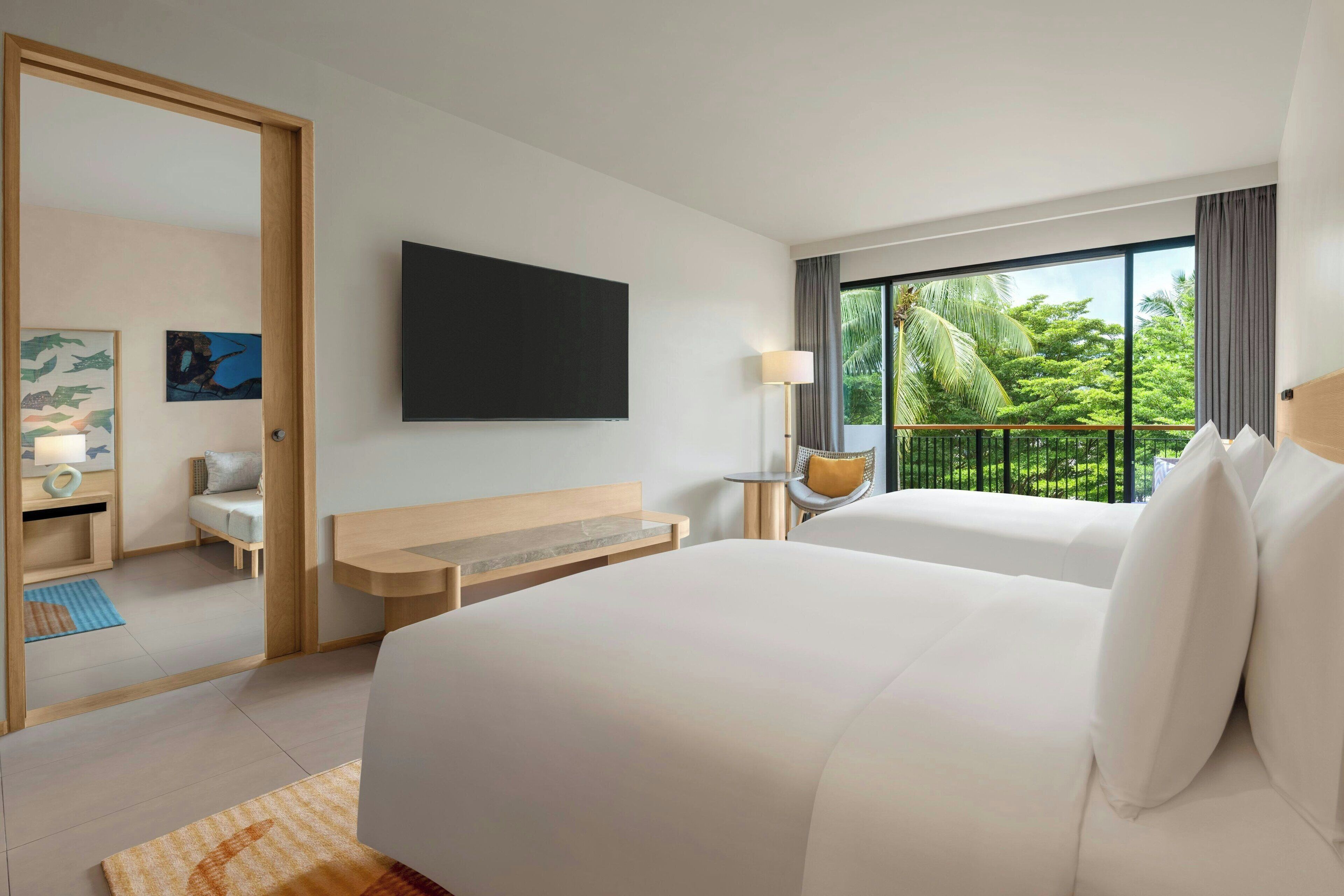 A hotel room with two beds, a desk, a chair, a TV, and a balcony with a view of greenery.