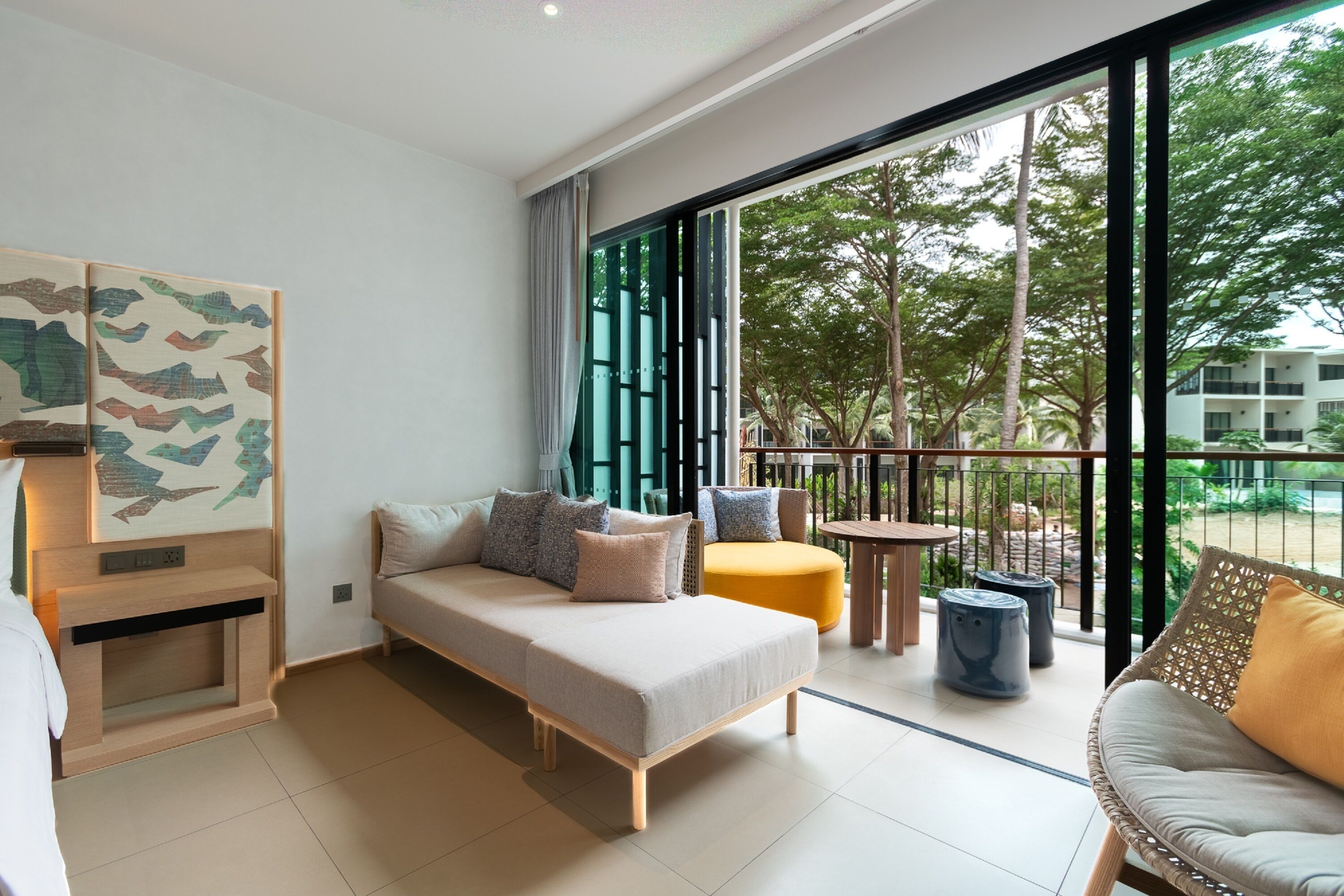 A modern hotel room with a sofa, a small table, and a balcony with a view of trees and buildings.
