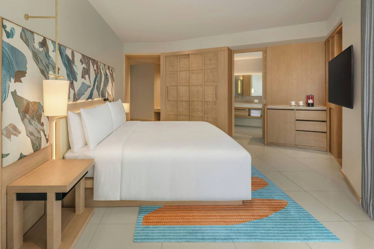 A hotel room with a bed, bedside table, lamp, artwork, wardrobe, and a television.