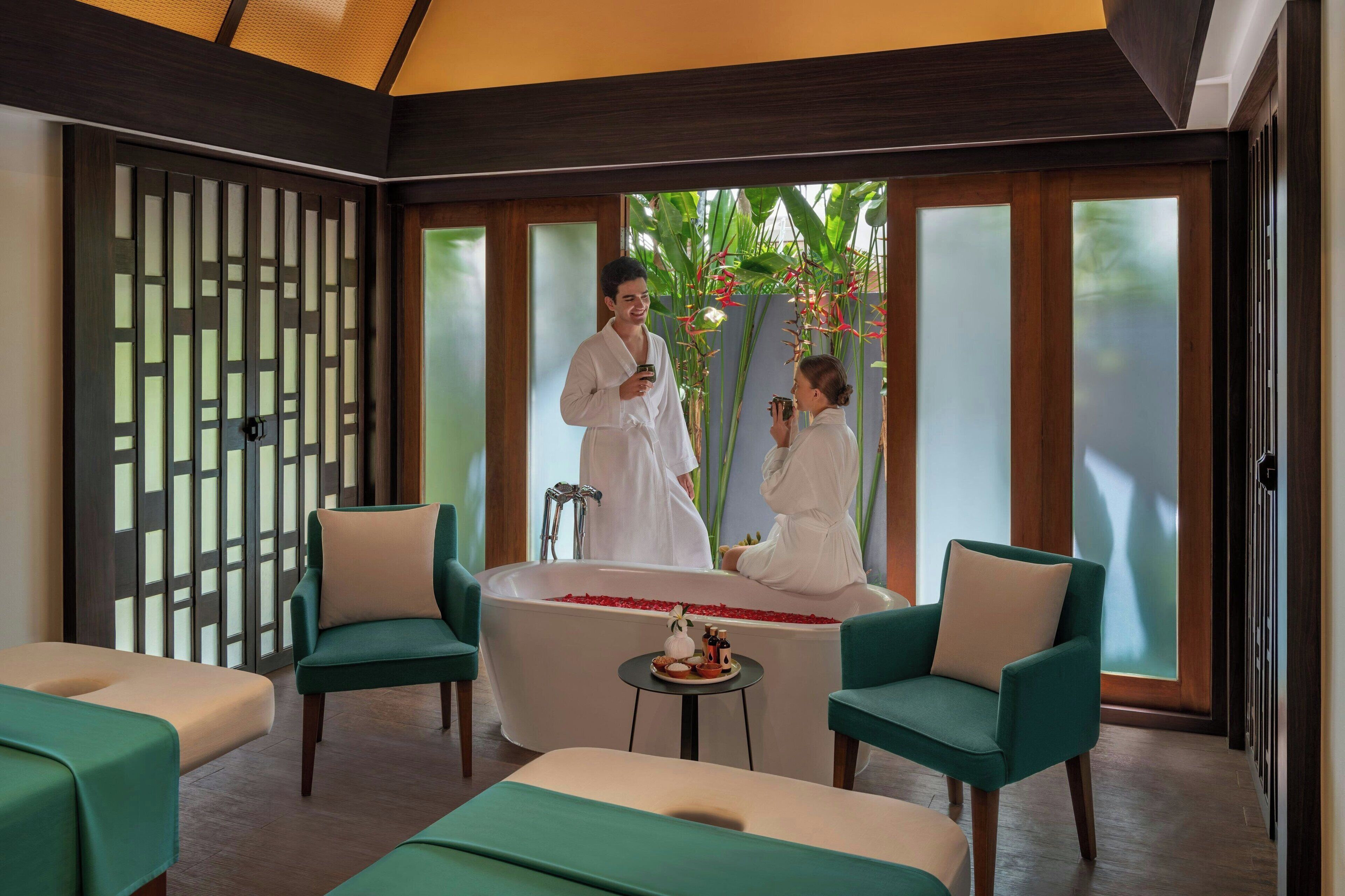 A spa room with a large bathtub, two green armchairs, and a small table with a decorative item.