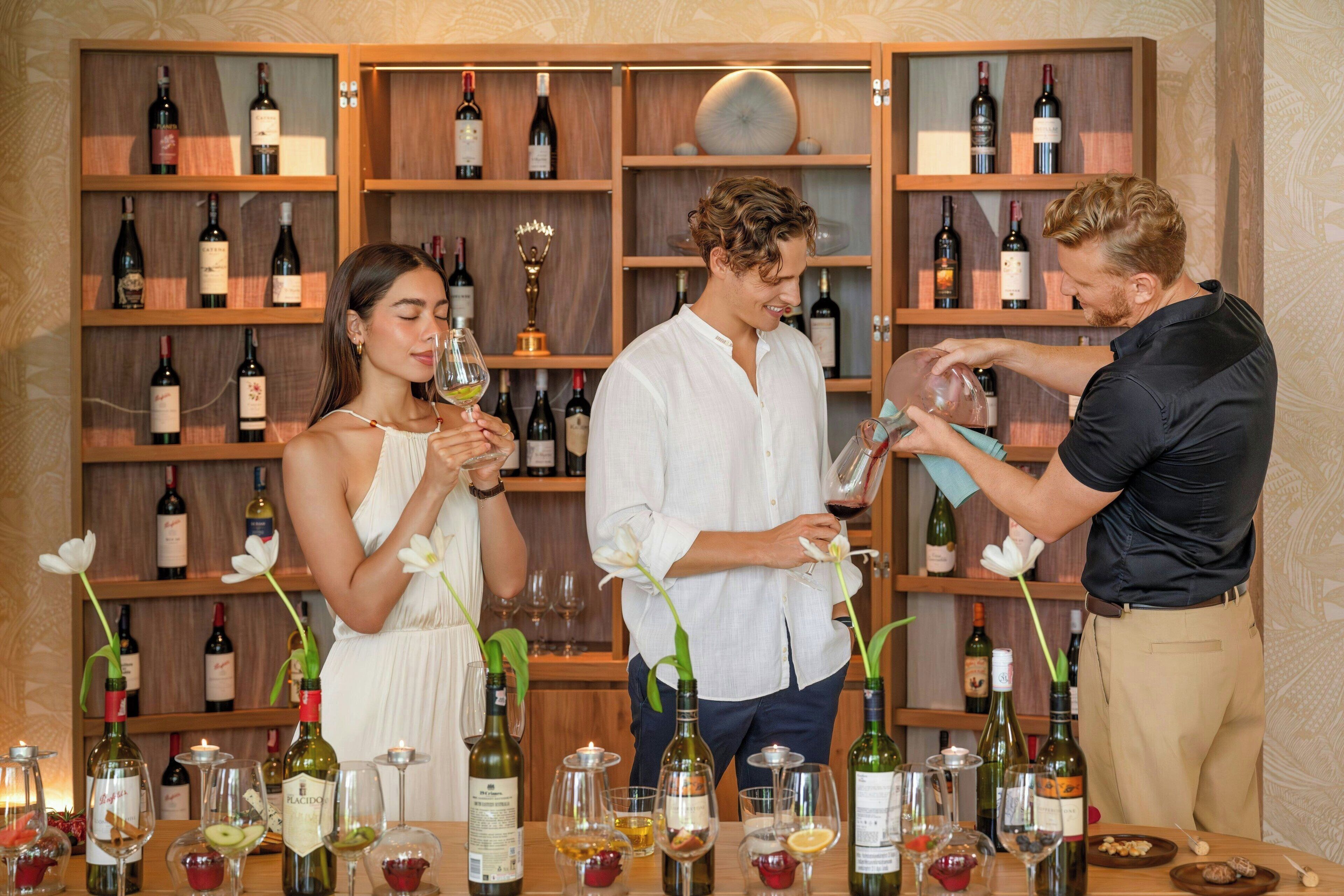 A bar area with a wooden wine rack in the background, a table with wine bottles and glasses, and a person pouring a drink.