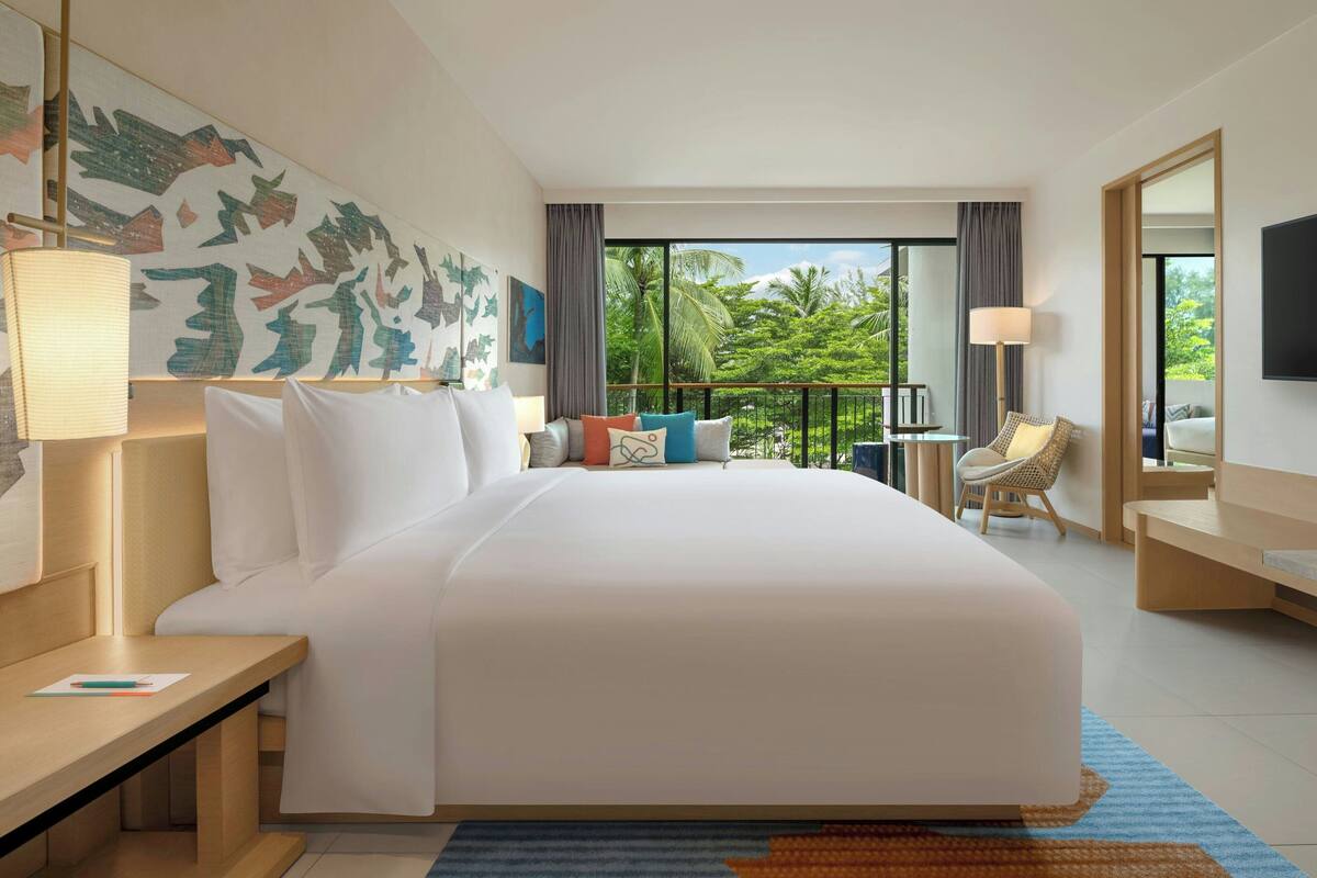 A hotel room with a large bed, a desk, a chair, a TV, and a balcony with a view of palm trees.