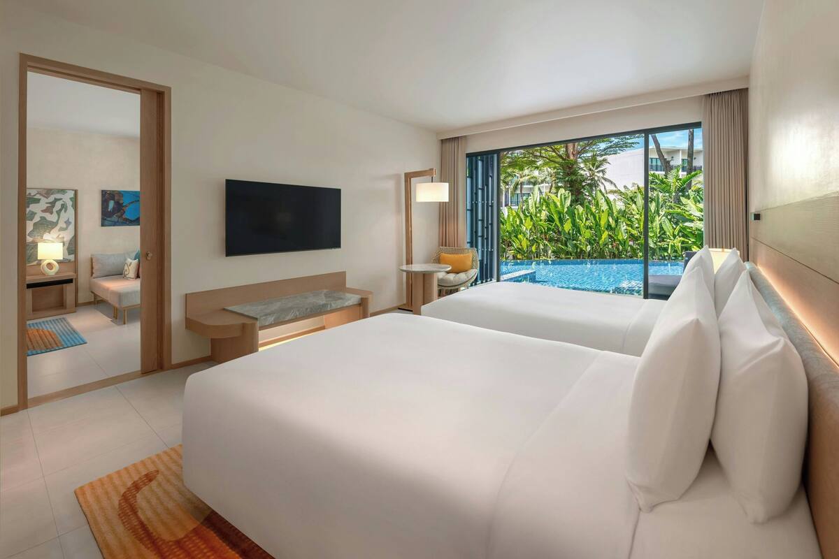 A hotel room with two beds, a TV, a small table with a lamp, and a view of a swimming pool.