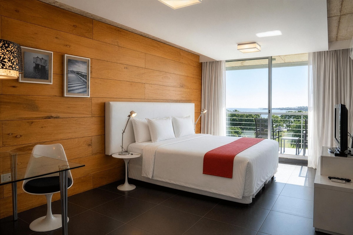A hotel room with a large bed, a desk, a chair, a nightstand, a TV, and a balcony with a view.