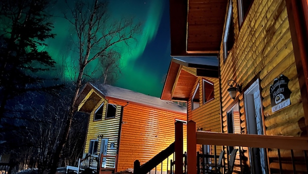 A log cabin with a staircase leading to the entrance, a sign indicating it is a Mountain Heather Chalet, and a view of the aurora borealis in the background. - Alaska Adventure Unlimited Chalets (Wasilla)
