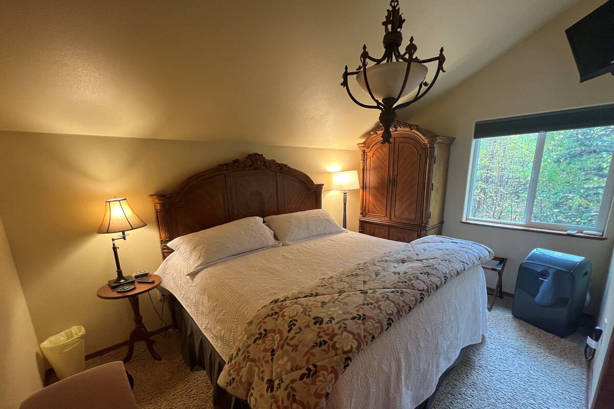 A bedroom with a large bed, two bedside lamps, a wooden wardrobe, a window with a view of trees, and a suitcase on the floor.