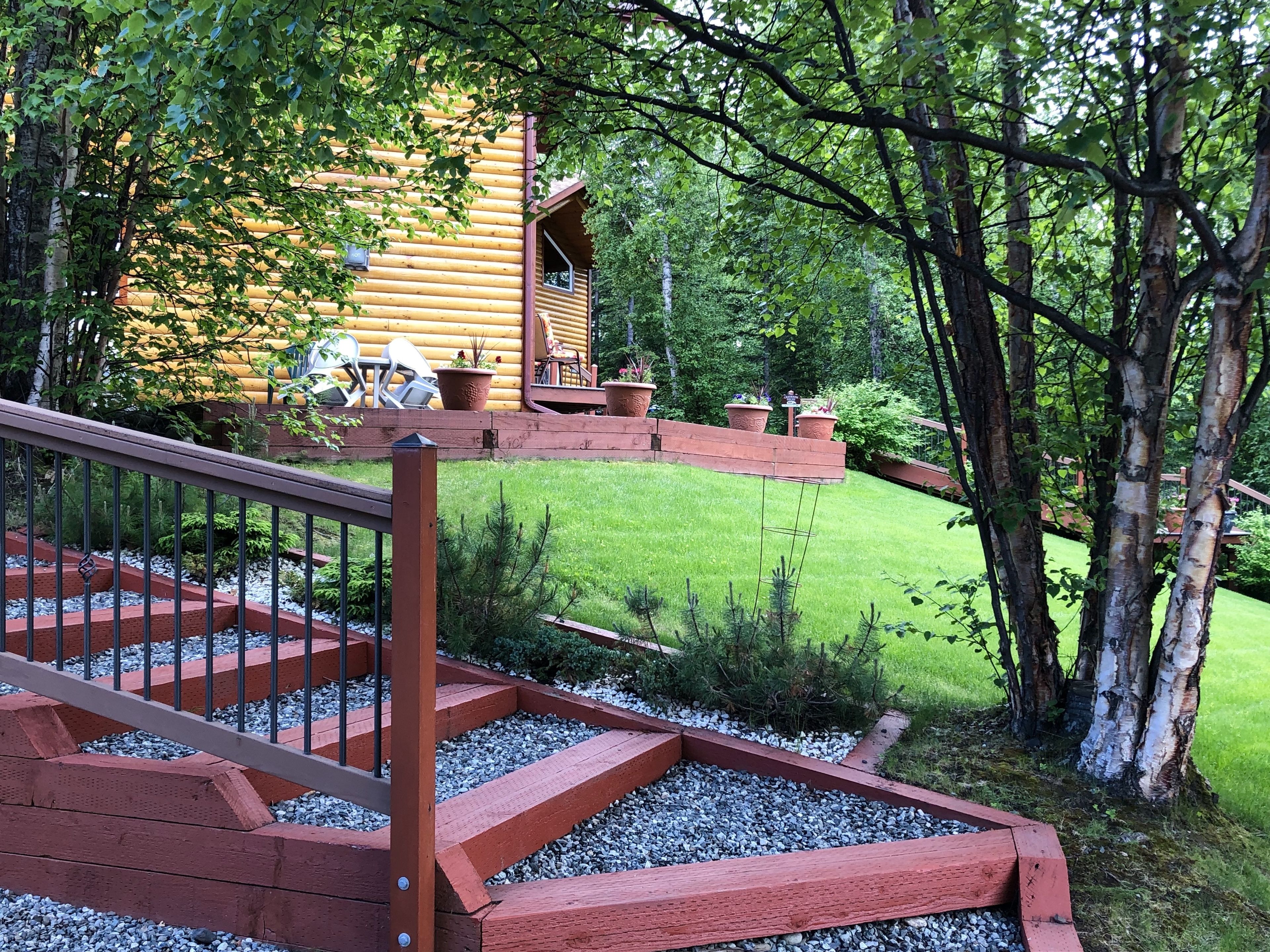 Une cabane en bois avec un escalier rouge menant à une terrasse, entourée d’une pelouse bien entretenue et d’arbres.