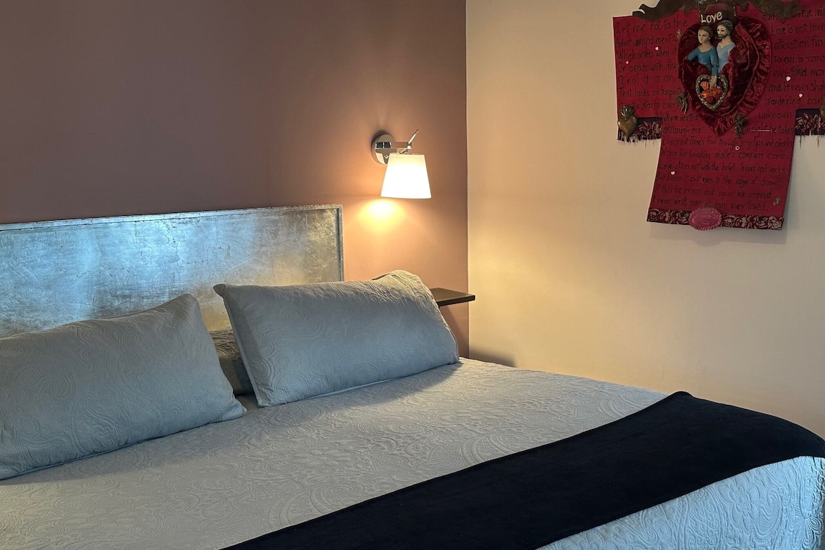 Una cama con una colcha estampada, dos almohadas y una manta de color negro. Hay una lámpara de pared y un objeto decorativo con texto e imágenes en la pared.