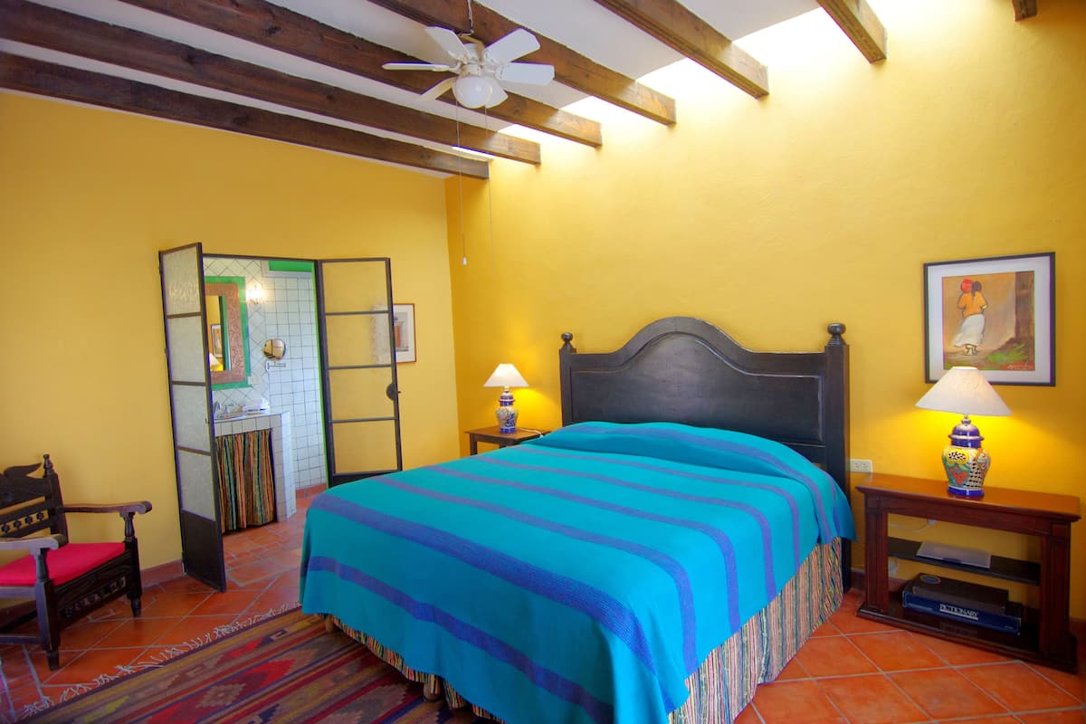Una habitación con cama, dos mesitas de noche, una silla, un espejo y un ventilador de techo.