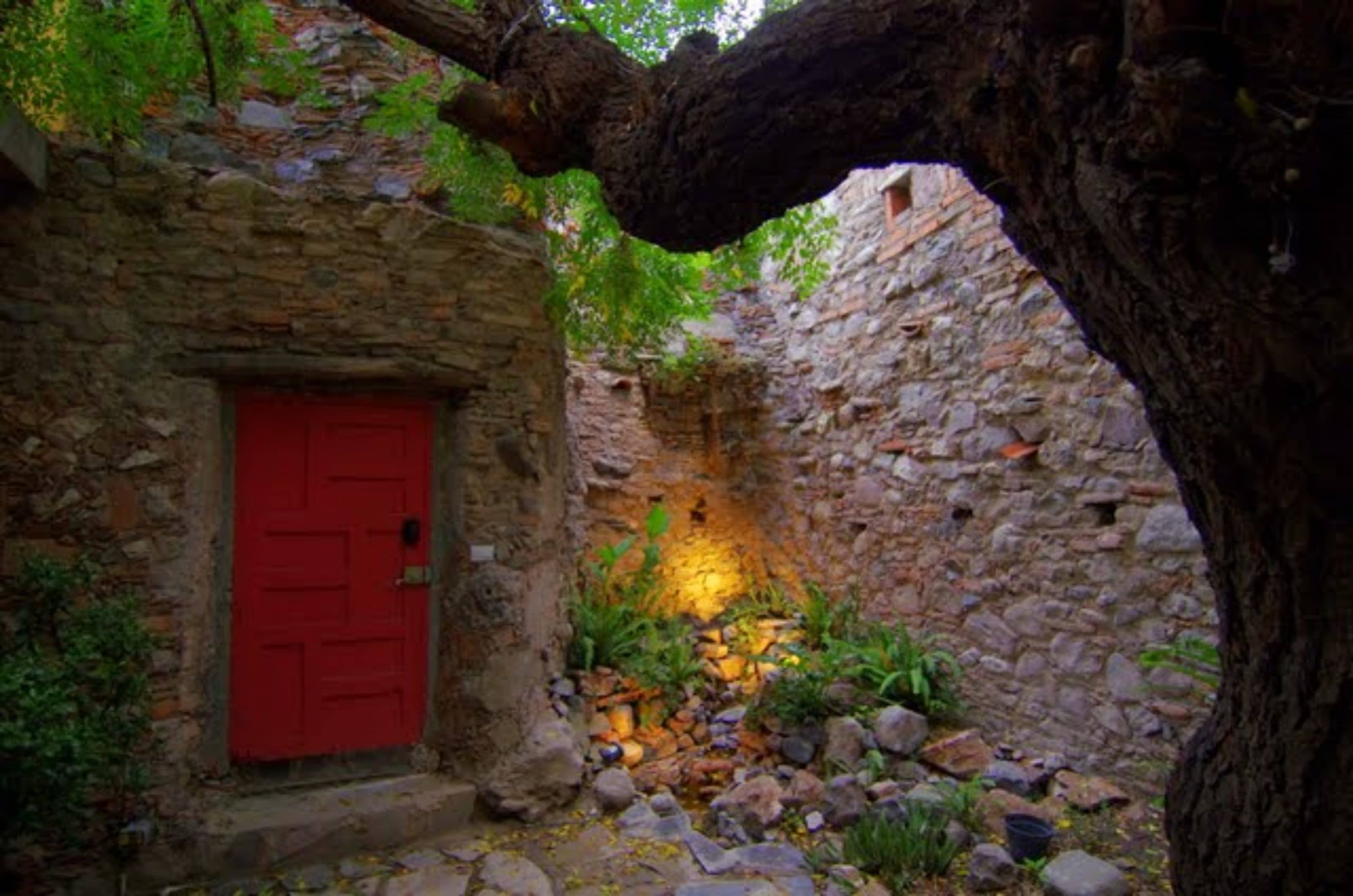 Un edificio de piedra con una puerta roja, un árbol y un pequeño jardín con rocas y plantas.