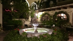 A courtyard with a central fountain, surrounded by greenery and potted plants. There are outdoor seating areas and a staircase leading to an upper level.
