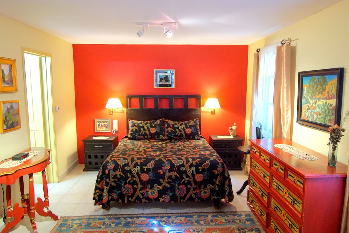 Un dormitorio con pared roja, cama con ropa de cama floral, dos lámparas de noche, una mesita de noche, un aparador y un cuadro enmarcado en la pared.