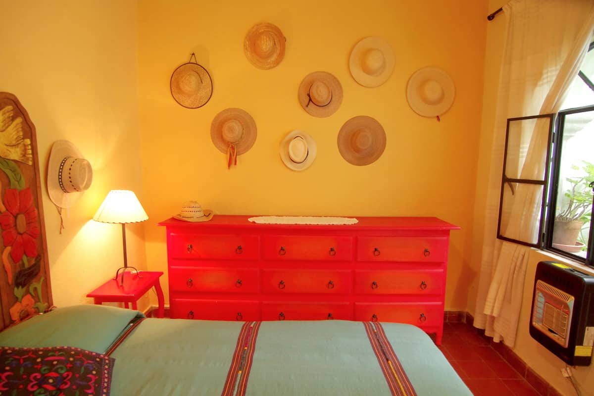 Una habitación con una cama, un aparador rojo, una pared con sombreros, una lámpara y una ventana con cortinas.