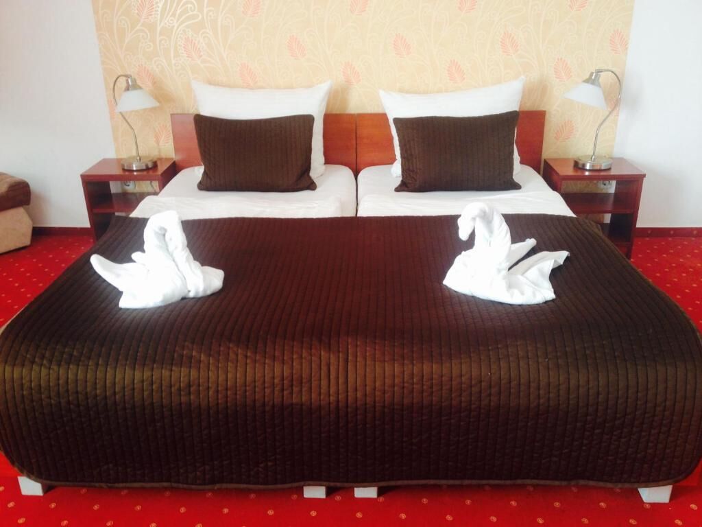 Una cama doble con una colcha marrón y dos toallas blancas con forma de cisne. Hay dos mesitas de noche con lámparas, un cabecero de madera y un papel tapiz estampado.