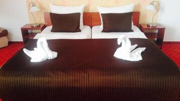 Una cama doble con una colcha marrón y dos toallas blancas con forma de cisne. Hay dos mesitas de noche con lámparas, un cabecero de madera y un papel tapiz estampado.