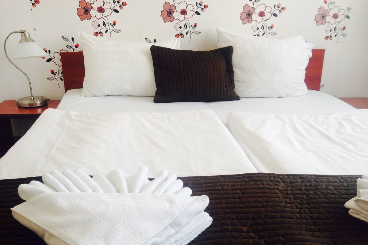 Una cama bien tendida con sábanas blancas y una manta marrón, una lámpara de noche y un estampado floral en el papel tapiz.