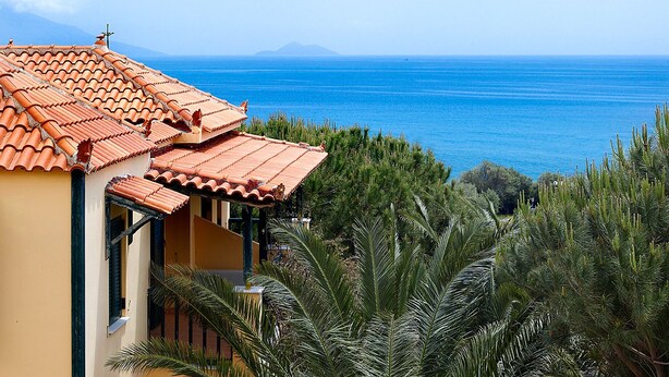 A coastal building with a tiled roof, surrounded by lush greenery and overlooking a clear blue sea.