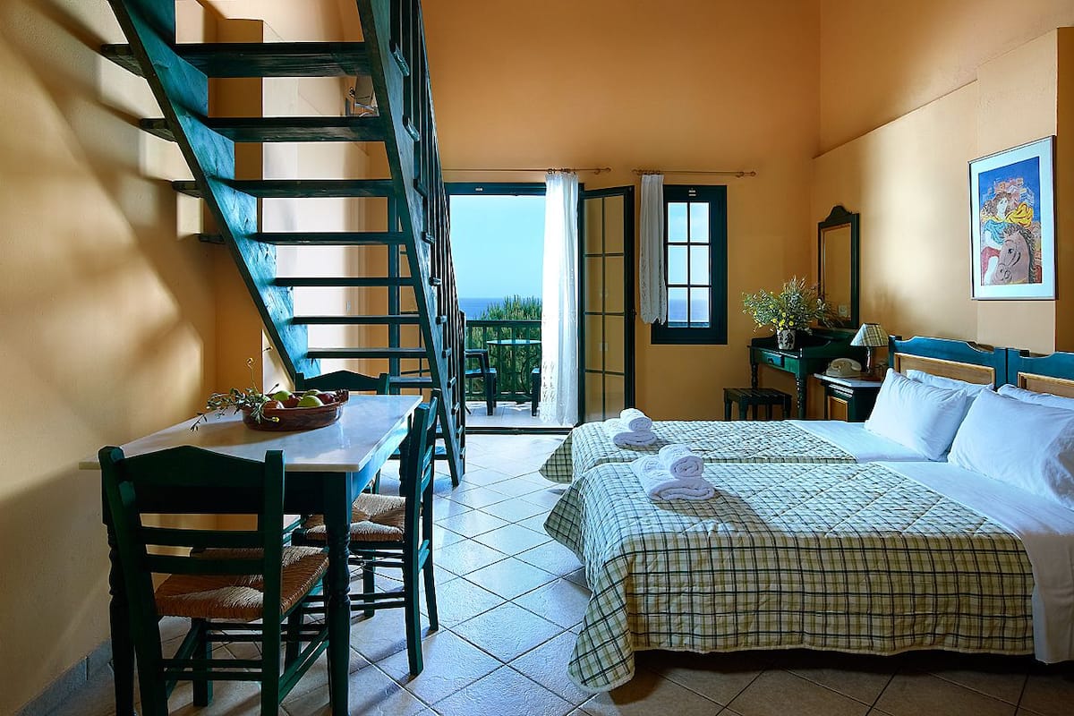 A bedroom with a bed, a table, chairs, a staircase, and a balcony with a view.