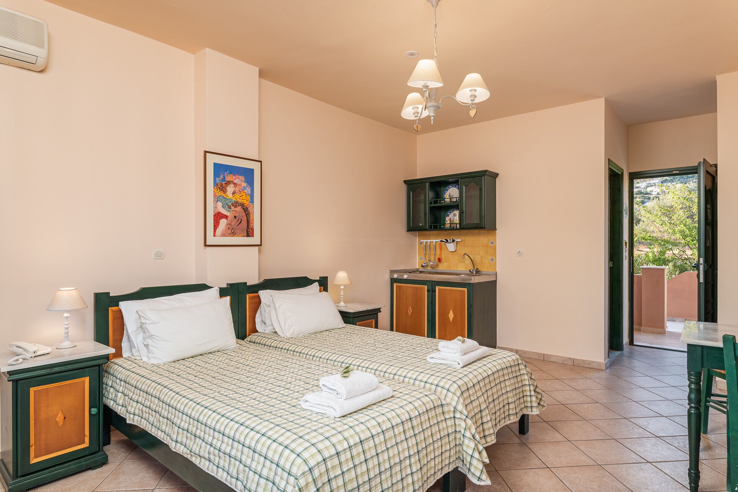 A hotel room with two beds, a kitchenette, a painting, and a view of the outside.