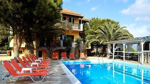 A swimming pool with red lounge chairs, a covered area, and a building with a tiled roof and balconies.