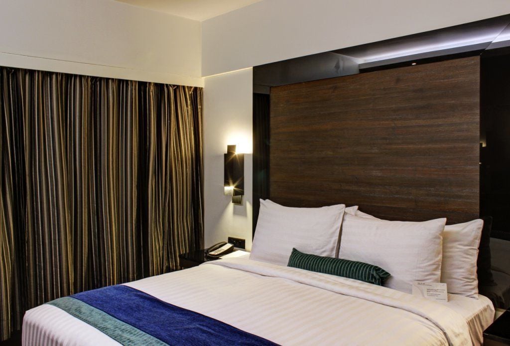 Una camera d'albergo con un letto grande, una testiera in legno, una tenda, un comodino con una lampada e un telefono.