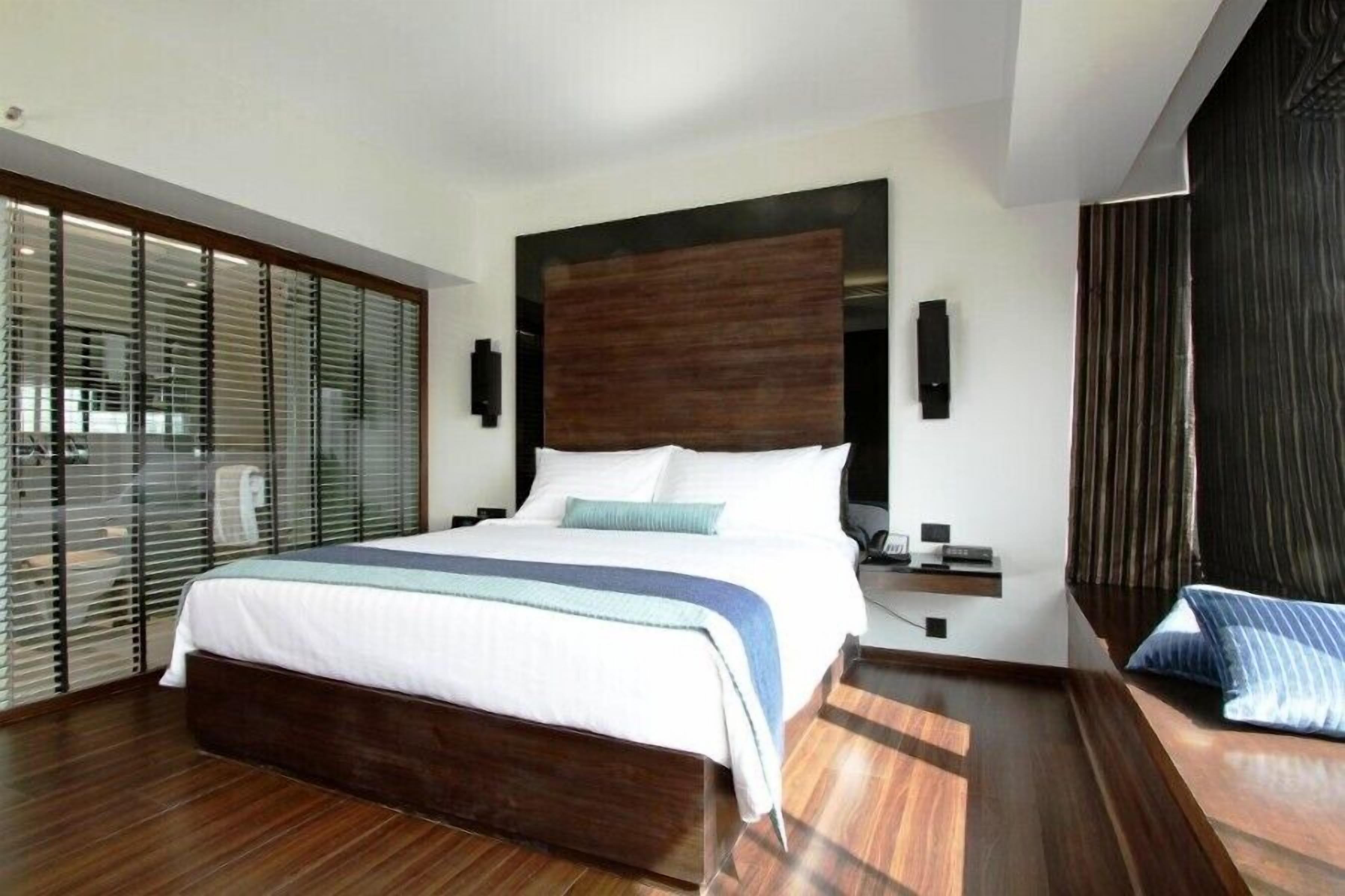A modern hotel room with a large bed, wooden flooring, a dark headboard, and a view through floor-to-ceiling windows.