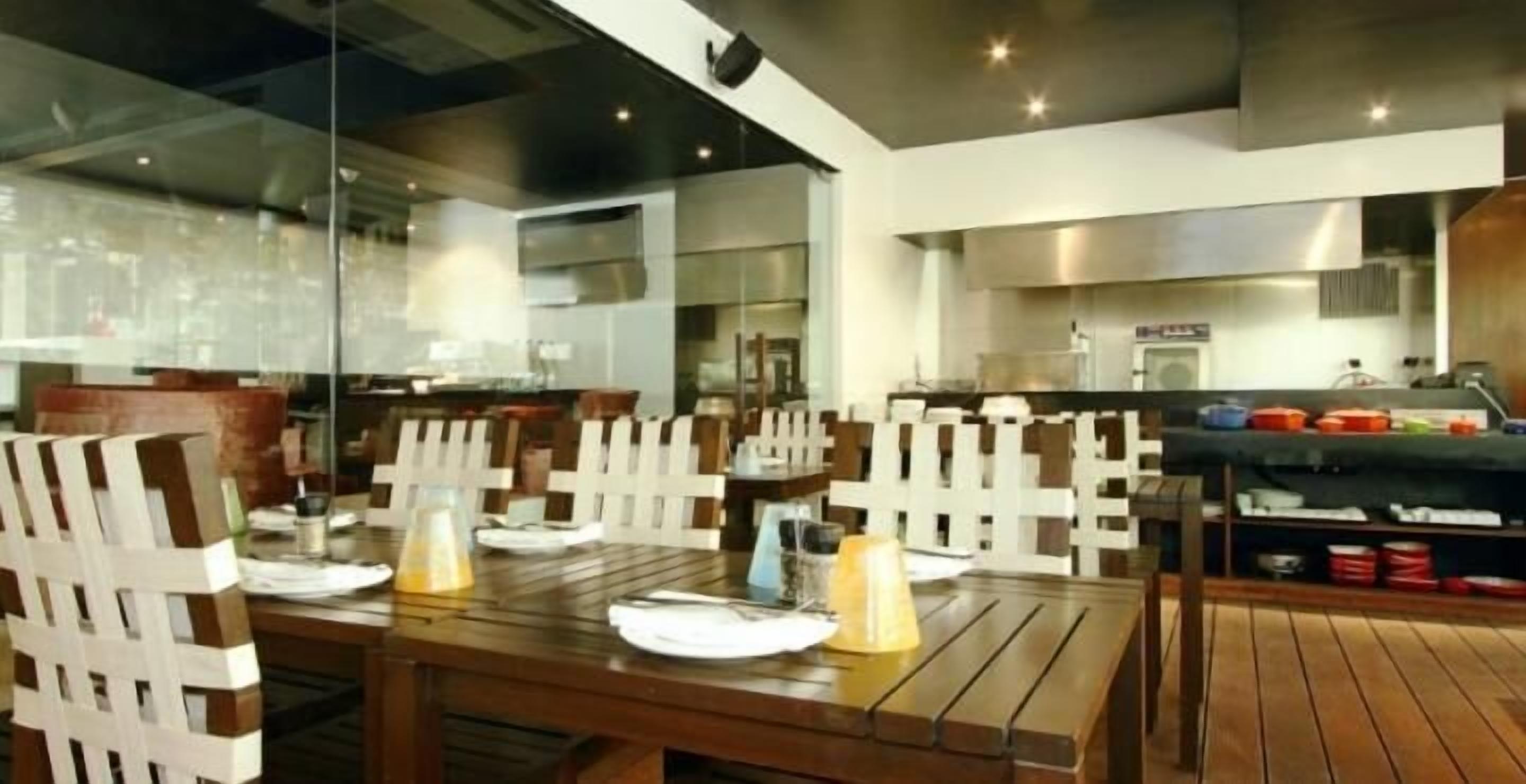 A restaurant with wooden tables and chairs, a glass partition, and a kitchen area in the background.