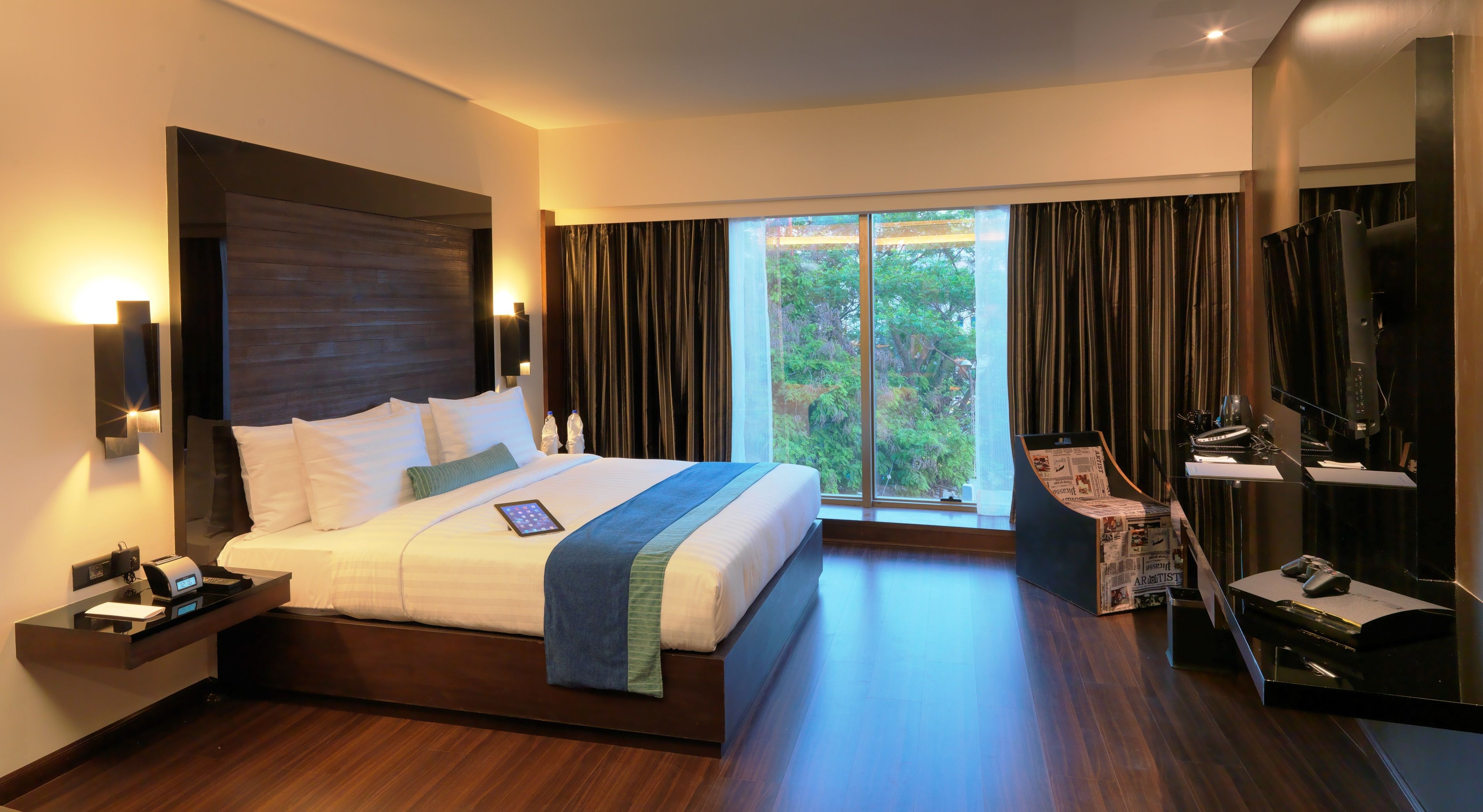 A modern hotel room with a large bed, a desk with a computer, and a view of the outdoors.