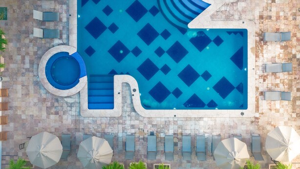 Vue aérienne d’une piscine dont le carrelage présente un motif géométrique, entourée de chaises longues et de parasols.