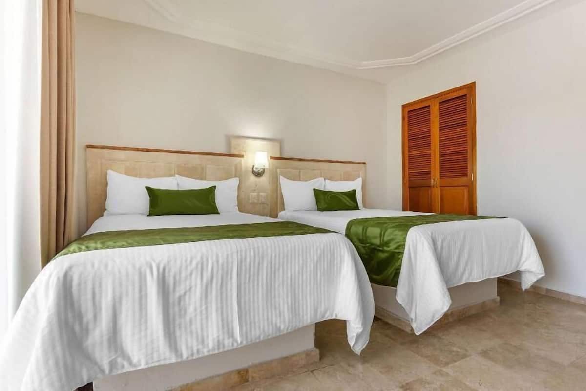 Habitación de hotel con dos camas, cabecera con lámpara y puerta de madera, sobre un suelo de baldosas color beige.