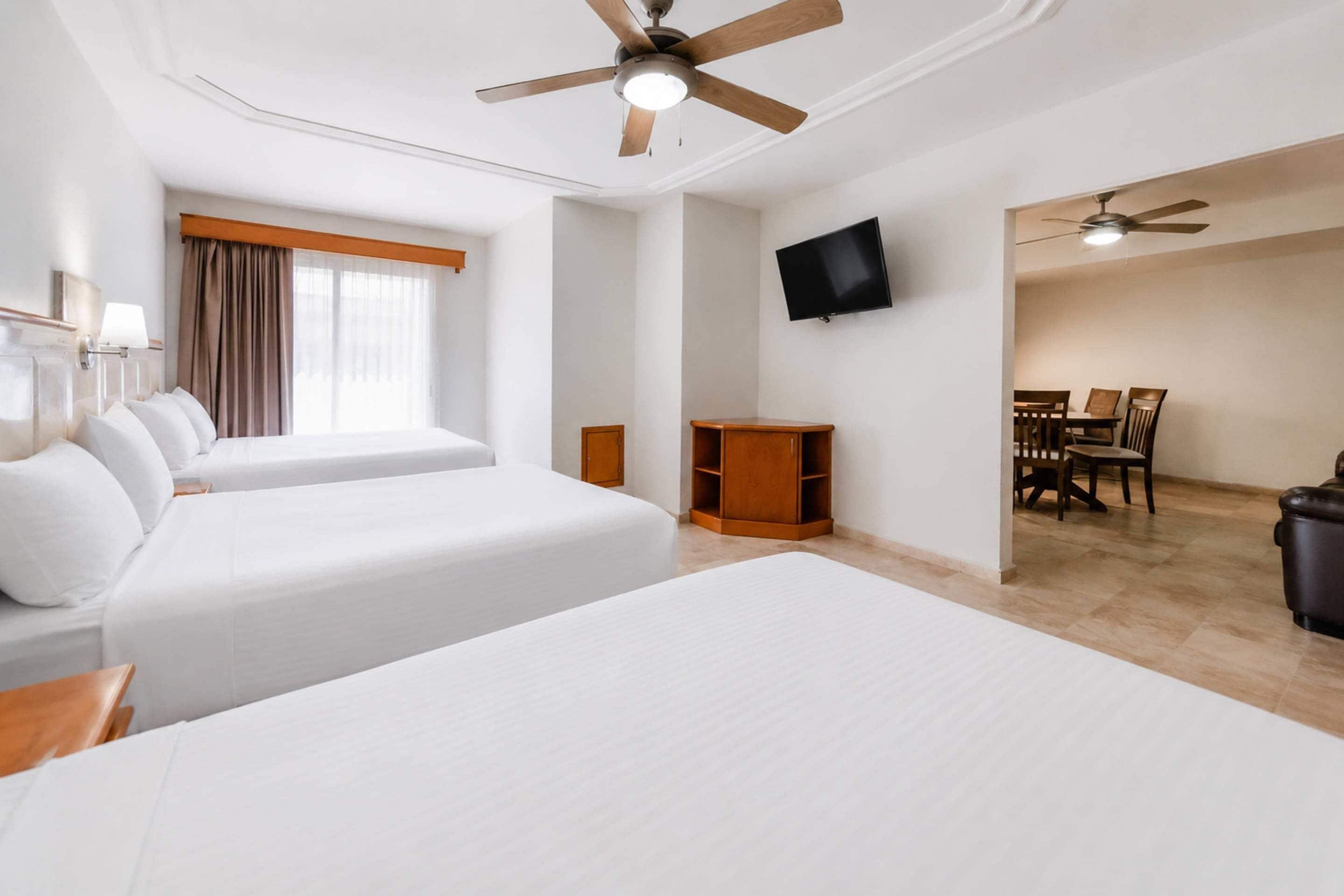 A hotel room with two beds, a TV, a ceiling fan, a dining area, and a trash bin.