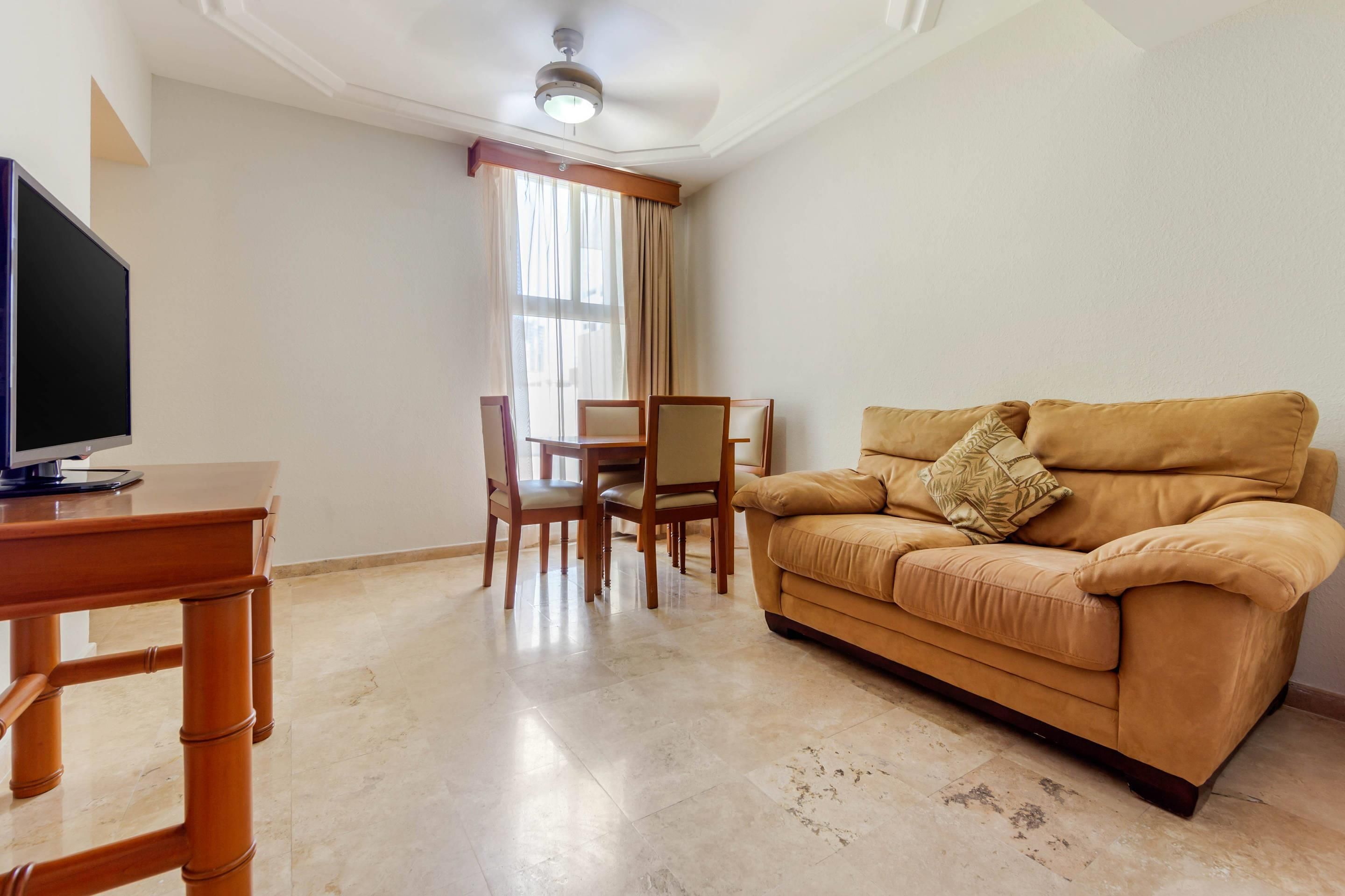A living room with a sofa, a TV on a wooden table, a dining table with chairs, and a window with curtains.