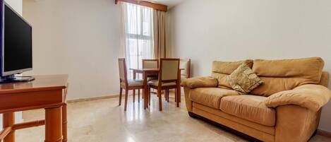 A living room with a sofa, a TV on a wooden table, a dining table with chairs, and a window with curtains.