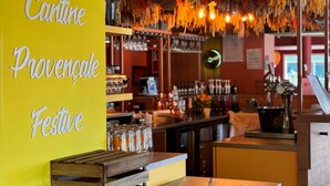 Zona de bar con estantes llenos de botellas y vasos, un cajón de madera y una pared amarilla con texto.