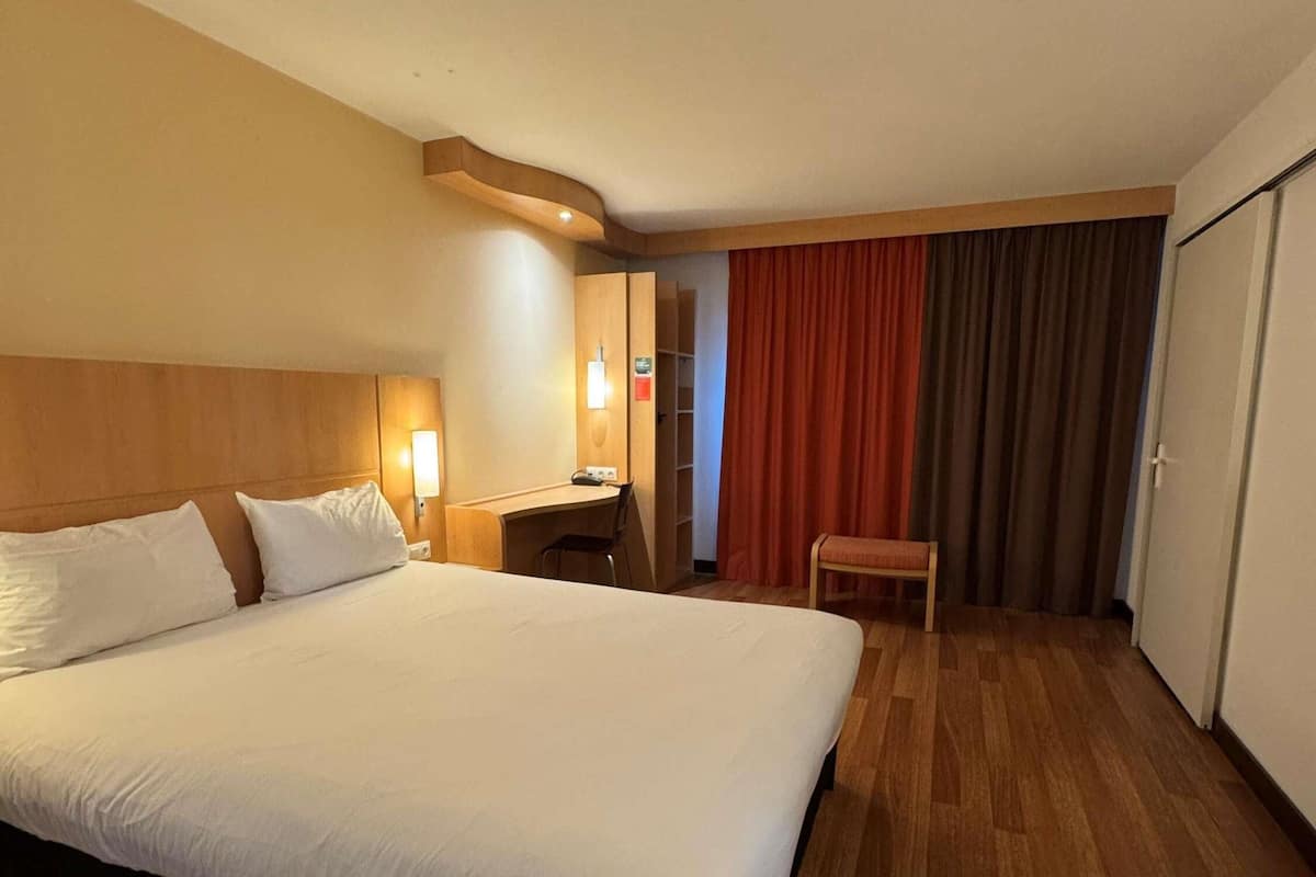 A hotel room with a large bed, a desk, a chair, a wardrobe, and a bathroom.