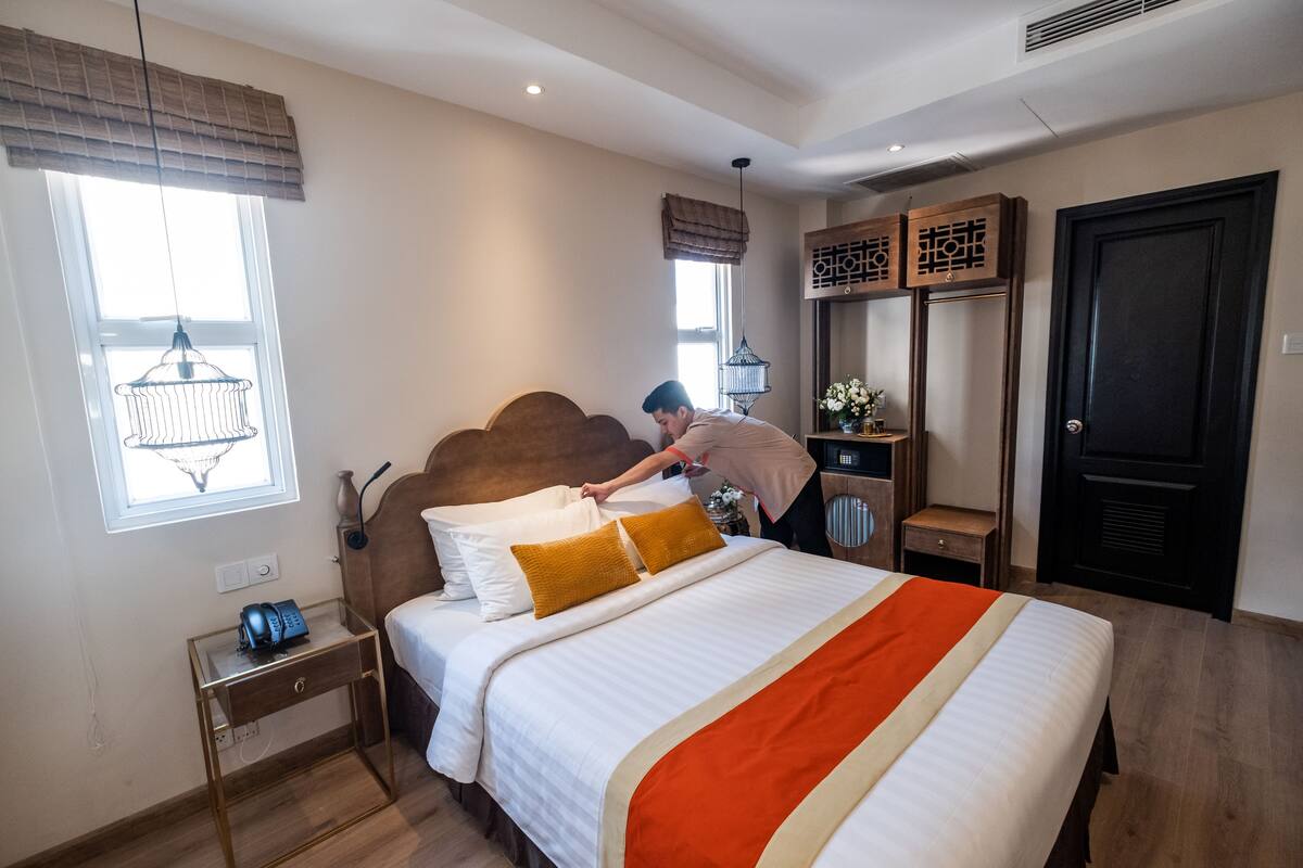 Un lit bien fait, recouvert d’une couverture blanche et orange, une table de chevet avec une lampe, et une fenêtre donnant sur l’extérieur.
