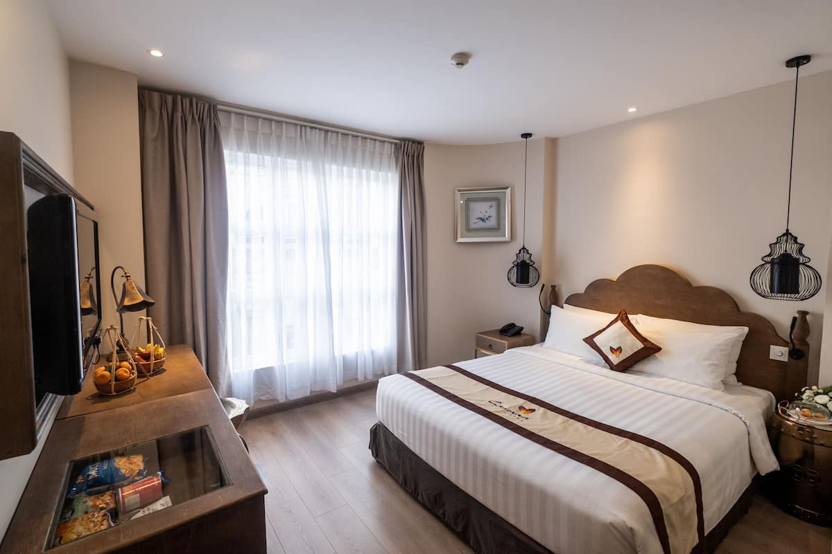 Une chambre d’hôtel avec un lit, une télévision, un bureau avec une lampe, une fenêtre avec des rideaux et un tableau encadré au mur.