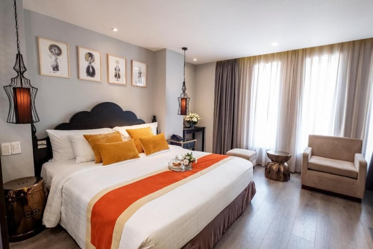 Une chambre d’hôtel avec un grand lit, un canapé, une table d’appoint et une chaise. Des tableaux encadrés ornent le mur et un lustre est suspendu au-dessus du lit.