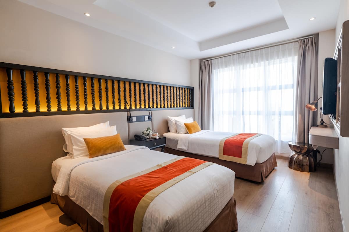 Une chambre d’hôtel avec deux lits, une grande fenêtre avec des rideaux, un luminaire fixé au mur et une petite table avec une lampe.